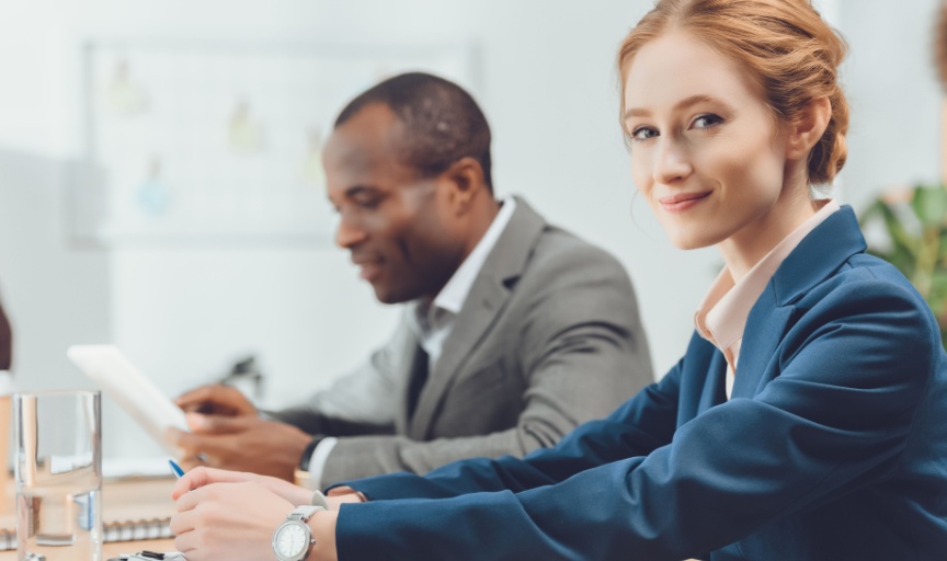 Two employees sit at their desks, with the staff member nearest looking straight at the camera, to illustrate the concept of employee experience