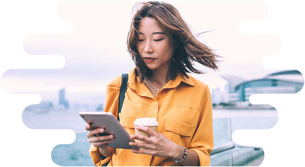 A female consumer standing outdoors completes an online questionnaire, using a tablet computer in one hand whilst holding a takeaway coffee in the other.