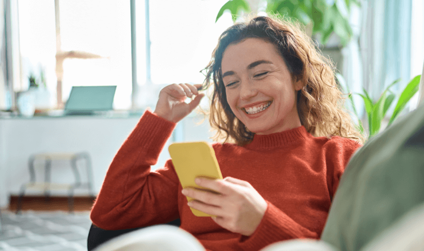 A female customer relaxes at home, expressing a positive sentiment by smiling at her smartphone